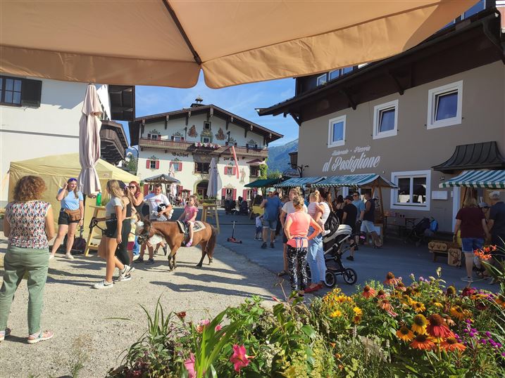 Ein lebendiger Marktplatz mit vielen Menschen und einem kleinen Pferd. Bunte Blumen und traditionelle Gebäude umrahmen die Szene.
