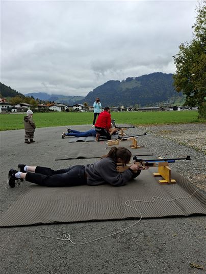 Mehrere Personen liegen auf Matten und üben mit Luftgewehren. Im Hintergrund sind eine grüne Landschaft und Berge zu sehen.
