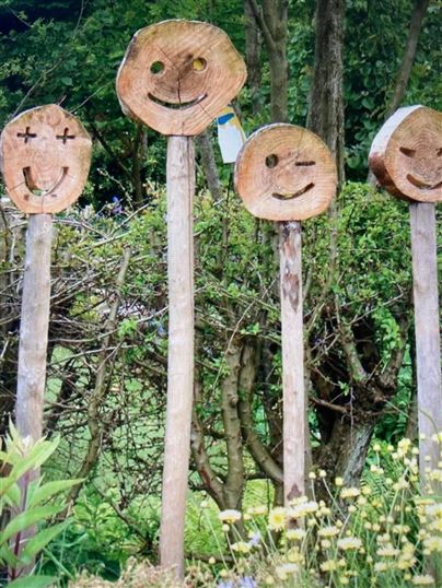 Vier Holzskulpturen mit fröhlichen Gesichtern stehen in einem Garten. Im Hintergrund sind grüne Pflanzen und Sträucher sichtbar.