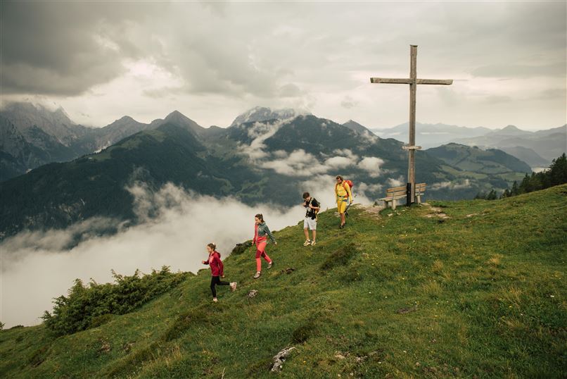 Eine Gruppe von Wanderern erkundet eine grüne Berglandschaft. Im Hintergrund sind Wolken und Gipfel zu sehen, sowie ein großes Kreuz.