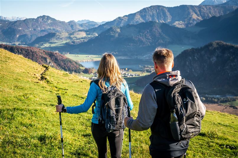 Zwei Wanderer stehen auf einer grünen Wiese und blicken auf eine wunderschöne Berglandschaft. Die Sonne scheint und der Himmel ist blau, während die Gipfel im Hintergrund deutlich sichtbar sind.