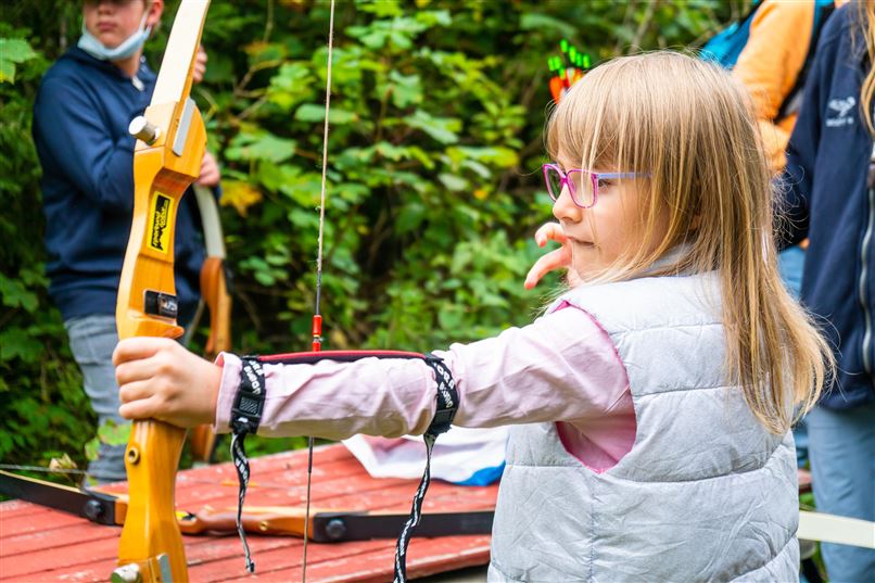 Ein Mädchen mit Brille zielt mit einem Bogen. Im Hintergrund sind weitere Personen und grüne Bäume zu sehen.