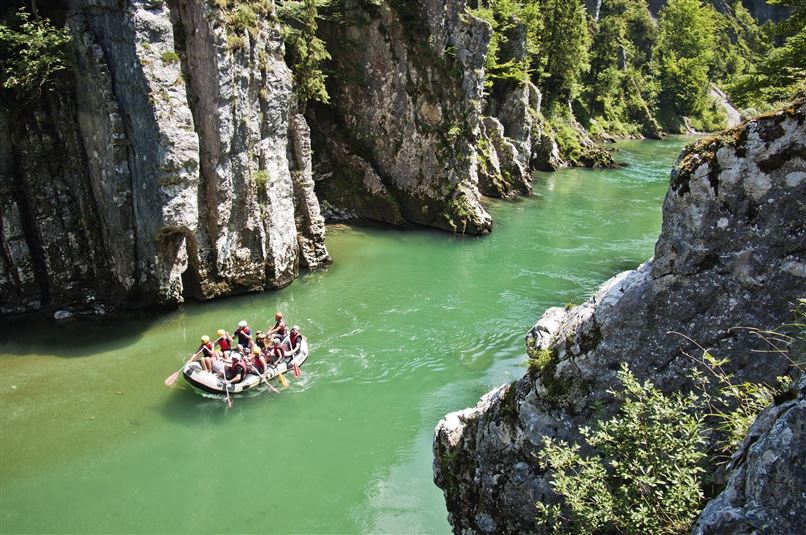 Eine Gruppe von Menschen paddelt in einem Schlauchboot auf einem klaren, grünen Fluss zwischen hohen Felsen. Die Umgebung ist grün und ruhige Natur.
