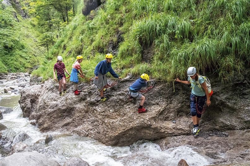 Eine Gruppe von Personen wandert entlang eines schmalen Pfades neben einem Fluss. Alle tragen Helme und scheinen den Weg in der Natur zu erkunden.