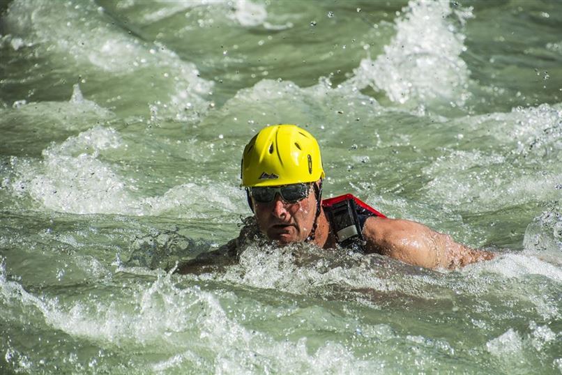 Ein Mann schwimmt kraftvoll in einem reißenden Fluss. Er trägt einen gelben Helm und eine Schwimmweste.