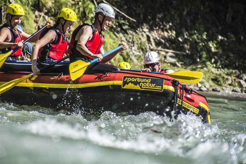 Eine Gruppe von Raftingfahrern paddelt in einem bunten Schlauchboot auf einem rauschenden Fluss. Alle tragen Helme und Schwimmwesten für Sicherheit.
