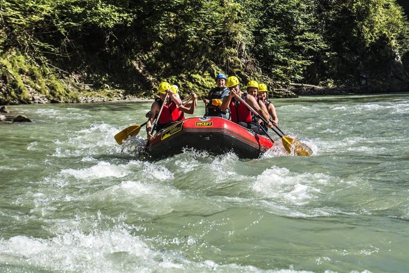Eine Gruppe von Personen paddelt in einem roten Rafting-Boot auf einem reißenden Fluss. Die Szene zeigt Spaß und Abenteuer inmitten einer grünen Umgebung.