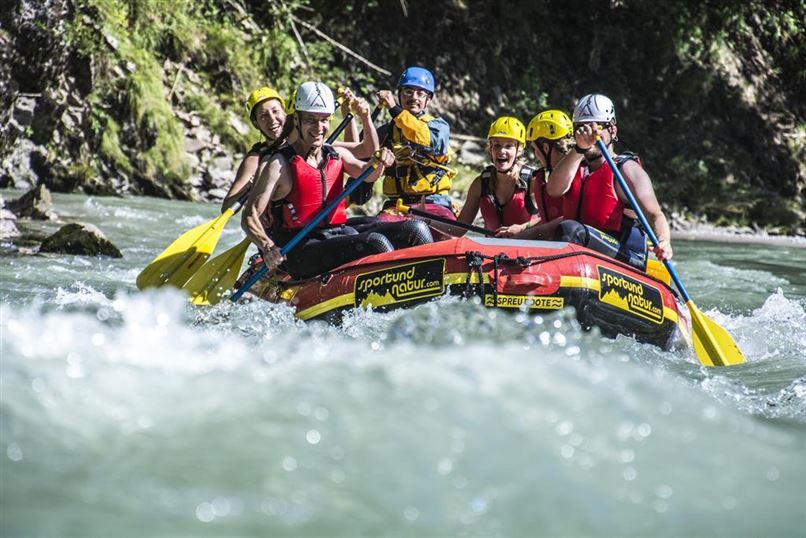 Eine Gruppe von sechs Personen paddelt in einem Raft auf einem wildem Fluss. Alle tragen Schwimmwesten und Helme und haben Spaß bei ihrem Abenteuer.
