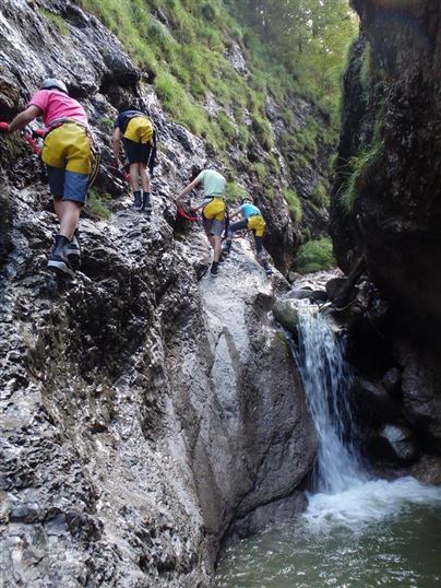 Gruppen von Menschen klettern an einer Felswand in einer Schlucht. Im Hintergrund fließt ein kleiner Wasserfall.