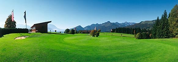 A green landscape with well-kept lawns and a beautiful view of the mountains. In the foreground stands a building with flags.