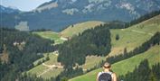 Zwei Wanderer gehen einen Hügel hinauf, umgeben von grünen Wiesen und Bergen im Hintergrund. Der Himmel ist klar und blau.