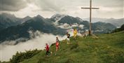 Eine Gruppe von Wanderern erkundet eine grüne Berglandschaft. Im Hintergrund sind Wolken und Gipfel zu sehen, sowie ein großes Kreuz.