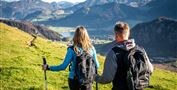 Zwei Wanderer stehen auf einer grünen Wiese und blicken auf eine wunderschöne Berglandschaft. Die Sonne scheint und der Himmel ist blau, während die Gipfel im Hintergrund deutlich sichtbar sind.