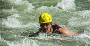 Ein Mann schwimmt kraftvoll in einem reißenden Fluss. Er trägt einen gelben Helm und eine Schwimmweste.
