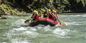 Eine Gruppe von Personen paddelt in einem roten Rafting-Boot auf einem reißenden Fluss. Die Szene zeigt Spaß und Abenteuer inmitten einer grünen Umgebung.