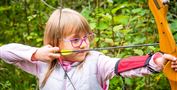 Ein Mädchen mit Brille zielt mit einem Bogen im Wald. Die Umgebung ist grün und naturbelassen.