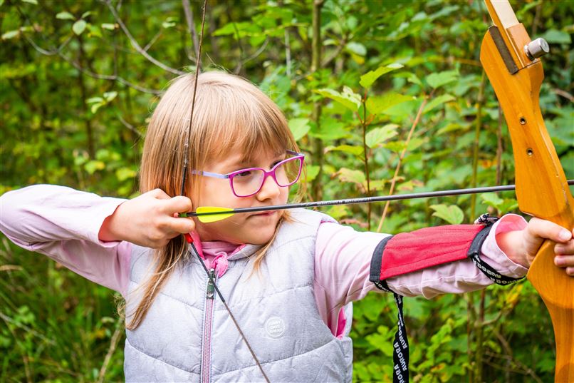 Ein Mädchen mit Brille zielt mit einem Bogen im Wald. Die Umgebung ist grün und naturbelassen.