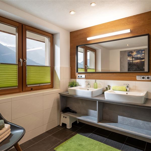Ein modernes Badezimmer mit zwei Waschbecken und einem großen Spiegel. Große Fenster lassen viel Licht herein und bieten einen Blick auf die Berge.
Weiters verfügt das Bad über 2 Duschen und einen riesenlangen Heizkörper welcher sich durch die Haken darüber im Winter perfekt eignet um nass gewordene Kleidung zu trocknen.