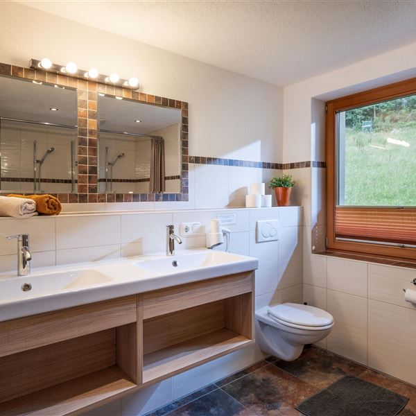 Ein modernes Badezimmer mit Doppelwaschbecken und großen Spiegeln. Viel Tageslicht strömt durch das Fenster, das einen Blick ins Grüne bietet.
Es bietet weiters eine Toilette und 2 Duschen damits schnell gehen kann.
