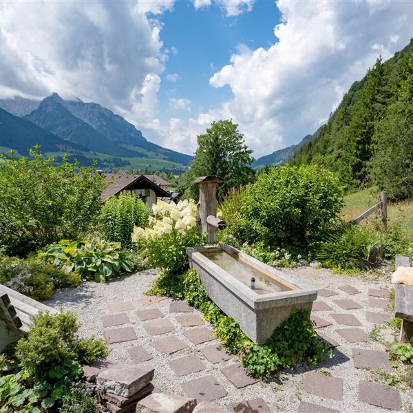 Ein malerischer Garten mit einem kleinen Brunnen und Bänken. Im Hintergrund erheben sich die Berge des Zahmen Kaisers an einem herrlichen Sommertag - Seeblick inklusive