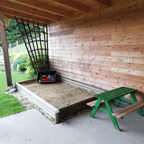 Eine Sandkiste mit Spielzeug steht in einem überdachten Bereich, egal ob die Sonne brennt oder der Regen fällt, ihre Kinder sind geschützt und haben immer Beschäftigung außerhab des Hauses. Daneben befindet sich ein kleiner Tisch und eine Bank.