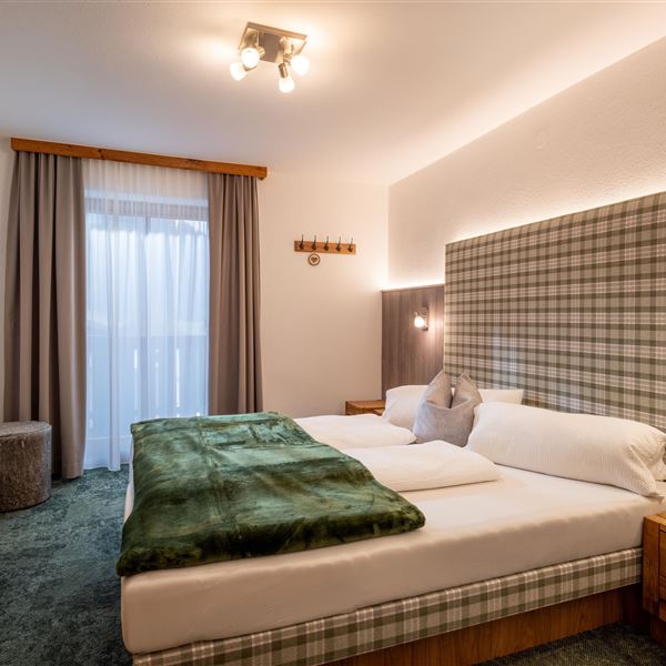Sie sehen hier  das erste Schlafzimmer im Obergeschoß, 2026 renoviert,  mit einem großen Bett (200x210) und einem karierten Kopfteil. Der Raum ist hell und gemütlich, mit einer Balkontür und Balkonzugang, Fliegengitter, Verdunkelungsvorhängen und  Eichenmöbeln.