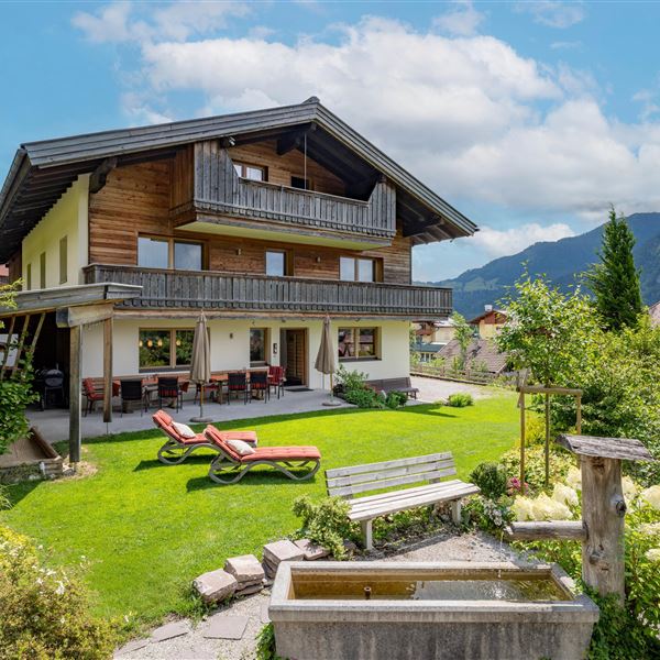 Ein modernes, großes Ferienhaus mit einem gepflegten Garten und entspannenden Liegen. Im Hintergrund sind Berge und ein blauer Himmel zu sehen. Im Vordergrund sehen Sie unseren Brunnen.