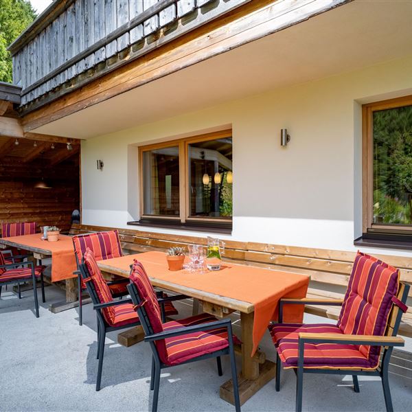 Eine einladende Terrasse mit großen Tischen und Stühlen in einem ruhigen Holzhaus. Der Sitzbereich auf der Terrasse umfasst 2 Bereich, die 2 Tische direkt vor dem Küchenfenster sowie ein weiterer Sitzbereich im überdachten Bereich etwas oberhalb. 
Die Umgebung ist grün und ruhig, ideal zum Entspannen.
